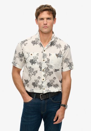 Camisa de mangas cortas de tela blanca con estampado floral negro, con cuello, dos bolsillos frontales y botones marrones. Combinada con vaqueros azules.