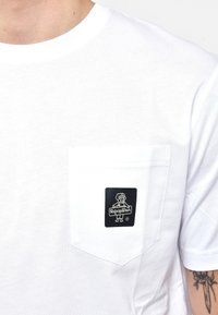 T-shirt bianco in cotone con una tasca sul petto e un'etichetta logo nera rettangolare che rappresenta un cane cartoon e la scritta "RefrigWear."