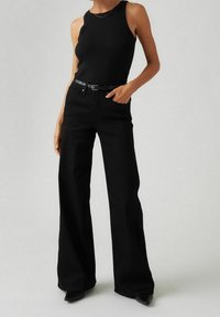 Personne portant un top sans manches noir, un pantalon large taille haute noir avec une ceinture, et des chaussures à talons noires sur un fond uni.