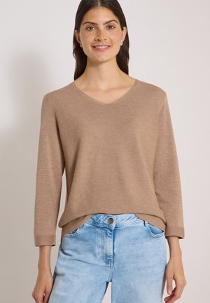 Femme aux longs cheveux foncés portant un pull à col en V marron clair et un jean taille haute bleu clair, souriante avec les mains le long du corps.