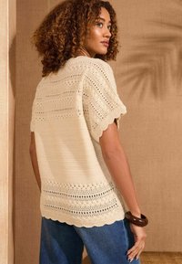 Maglione in cotone lavorato a maglia di colore crema con dettagli a rete aperta, bordi scallop e maniche corte, abbinato a pantaloni in denim blu. Sfondo semplice.