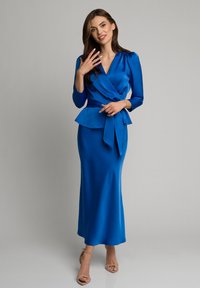 Niebieska satynowa sukienka o kroju kopertowym z peplum w pasie, długimi rękawami i paskiem do wiązania. Elegancka sylwetka o gładkiej fakturze.