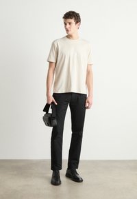 Beige cotton t-shirt with short sleeves and logo, paired with black trousers, and black lace-up shoes. Holding a small black bag.