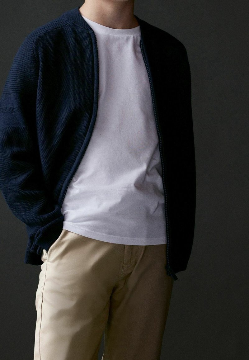 Personne portant un pull zippé bleu marine par-dessus un t-shirt blanc et un pantalon beige, debout avec une main dans la poche, devant un fond sombre.