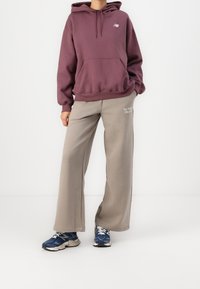 Sudadera con capucha color marrón y bolsillo frontal, combinada con pantalones de chándal de pierna ancha grises con logotipos blancos, y completada con zapatillas azules.