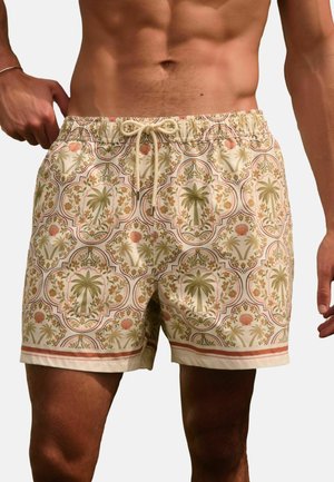 Muž v béžových plavkách s motivem palmy a květin, upravuje šňůrku na pasu.