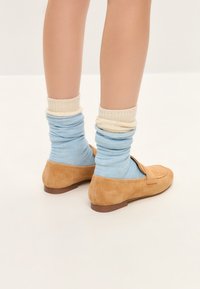 Barna suede loafer cipők, kék és krém árnyalatú felgöngyölt zoknikkal párosítva, sima textúrával és egyszerű dizájnnal. A lábak enyhén szétfeszítve.