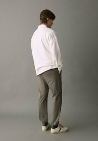 Man i vit långärmad skjorta, grå joggbyxor, vita sneakers med gröna ränder, stående med händerna i fickorna mot en enkel grå bakgrund.