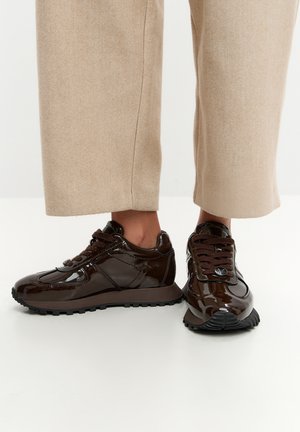 Osoba nosząca błyszczące ciemnobrązowe sneakersy na sznurowadła i beżowe spodnie z szerokimi nogawkami na białym tle.