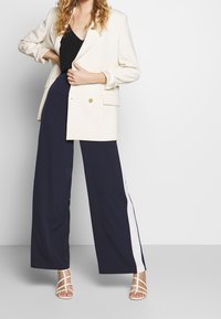 Krämfärgad dubbelknäppt kavaj med två framfickor, parat med marinblå vida byxor med en vit sidstripe och vita sandaletter med remmar.