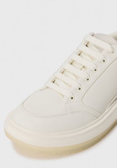 Fehér sneaker, sima bőrből készült felsőrésszel, kerek orral, hat fűzőlyukkal és áttetsző szélű, mintázott gumitalppal.