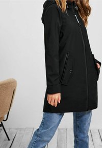 Černá nepromokavá bunda s kapucí, na zip, se dvěma bočními kapsami a nastavitelnými manžetami, v kombinaci s modrými džínami. Hladká textura.