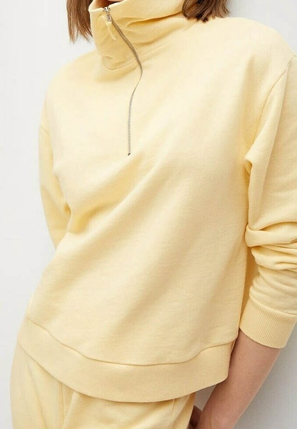 Femme portant un pull jaune clair à col montant avec demi-fermeture éclair, assorti à un pantalon, debout devant un fond blanc.