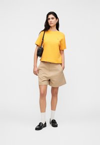 Κίτρινο βαμβακερό t-shirt με κοντά μανίκια, μπεζ σορτς, μαύρα loafers και λευκές κάλτσες. Μικρό μαύρο τσαντάκι ώμου. Μινιμαλιστικός σχεδιασμός, άνετη εφαρμογή.