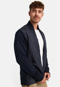 Navy bomber jacket with a zip front, knit sleeves, and textured fabric. Features ribbed cuffs and hem, layered with a white T-shirt underneath.