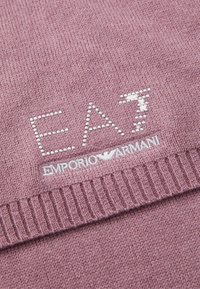 Rózsaszín kötött anyag ezüst gyöngy díszítéssel, amely az "EA7" feliratot alkotja a fehér hímezett "Emporio Armani" logó felett.