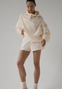 Jasno beżowa, lekka kurtka z wysokim kołnierzem i regulowanym sznurkiem, zestawiona z dopasowanymi szortami o gładkiej fakturze.