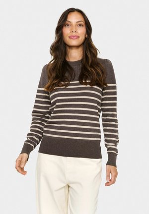 Kvinde med langt, bølget hår iført en brun og beige stribet sweater og hvide bukser, der står op ad en ensfarvet lys baggrund.