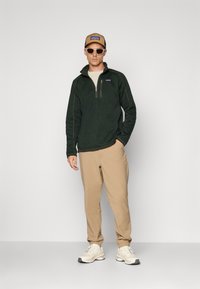 Grønn fleece genser med halv glidelås, beige bukser, beige caps med logo, og hvite solbriller. Skoene er kremfargede med grå detaljer.
