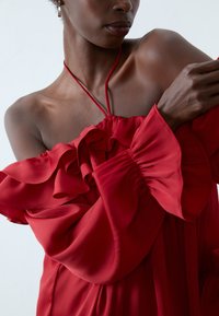 Robe rouge sans épaule avec des manches à volants et un corsage froncé. Fabriquée dans un tissu léger et texturé avec un design fluide.