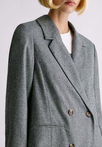 Donna con un bob biondo che indossa un blazer doppiopetto grigio in motivo a spina di pesce con rever e bottoni marroni sopra una maglietta bianca.