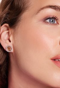 Gouden ronde oorbellen met een bloemmotief in het midden, omringd door een ring van kleine heldere kristallen, gedragen aan het oor van een model.