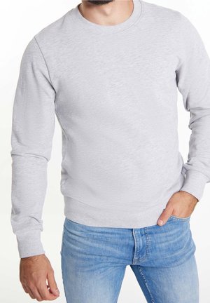 Homme portant un sweat-shirt gris clair à col rond et un jean bleu clair, main dans la poche, debout devant un fond blanc.