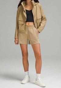 Femme portant une veste à capuche beige, un short assorti, un haut court noir, des chaussettes blanches et des baskets blanches, debout devant un fond gris.