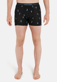 Manliga ben i svarta boxerkalsonger med ljusblå vinteridrottsymboler och "icebreaker" på midjebandet, stående barfota mot en vit bakgrund.