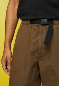 Personne portant un pantalon marron avec une ceinture en tissu noire et une chemise noire, main détendue sur le côté devant un fond jaune.
