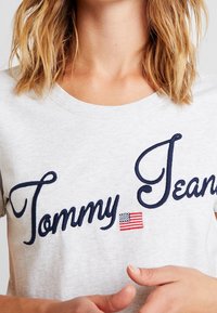Šedé bavlněné tričko s námořnicky modrým vyšívaným nápisem "Tommy Jeans" a malou vyšívanou americkou vlajkou. Kulatý výstřih.