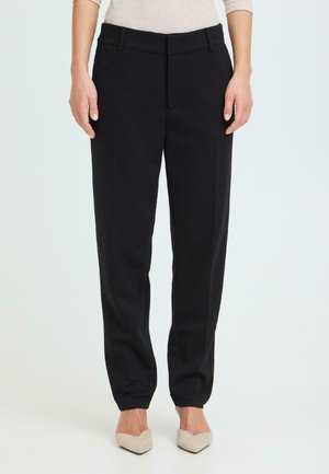 Kobieta ubrana w czarne, dopasowane spodnie z podwiniętymi nogawkami i beżowe szpilki z ostrym noskiem, stojąca na tle jasnego, jednolitego tła.