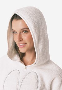 Biała, puszysta odzież z kapturem, posiadająca zaokrąglony kołnierz, zapięcie na guziki oraz nadruk z przodu w postaci dwóch dużych, zaokrąglonych kształtów w kontrastowym kolorze.