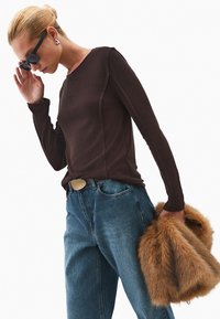 Bruine langarmshirt met een aansluitende snit, gecombineerd met comfortabele blauwe jeans en een accessoire met bontaccent. Donkere zonnebril maakt de look compleet.