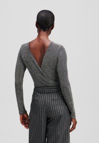 Garā piedurkņu apģērbs tumši pelēkā krāsā ar V-veida aizmuguri, ar teksturētu adījumu un izkaisītām metāliskām akcentiem, saderīgs ar pinstrīpu biksēm.