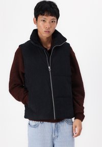Zwarte corduroy vest met hoge kraag en rits, gedragen over een donkerbruin gestructureerd trui, gecombineerd met lichtblauwe jeans.