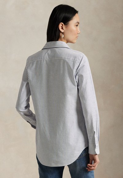 Kobieta z czarnymi włosami nosi białą koszulę z długim rękawem z cienkimi niebieskimi pionowymi paskami i niebieskie dżinsy, stojąc bokiem na beżowym tle.