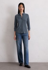 Hemd mit Knopfleiste aus dunkelblauem Stoff mit geometrischem Muster, langärmlig und hochgekrempelt, kombiniert mit hellblauen Jeans mit geradem Bein und schwarzen Schuhen.