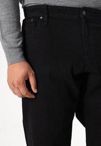 Blugi din denim negru cu o croială slim, având fermoar și nasture, buzunare frontale și o textură fină. Mână odihnindu-se pe partea laterală.