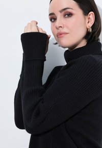 Maglione girocollo a costine nere con maniche allungate. Texture liscia, design aderente e collo alto. Orecchino a cerchio dorato visibile.