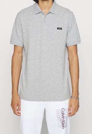 Mężczyzna nosi jasnoszary polo z krótkim rękawem z małym czarnym logo Calvin Klein oraz białe spodnie z napisem Calvin Klein na lewym udzie.