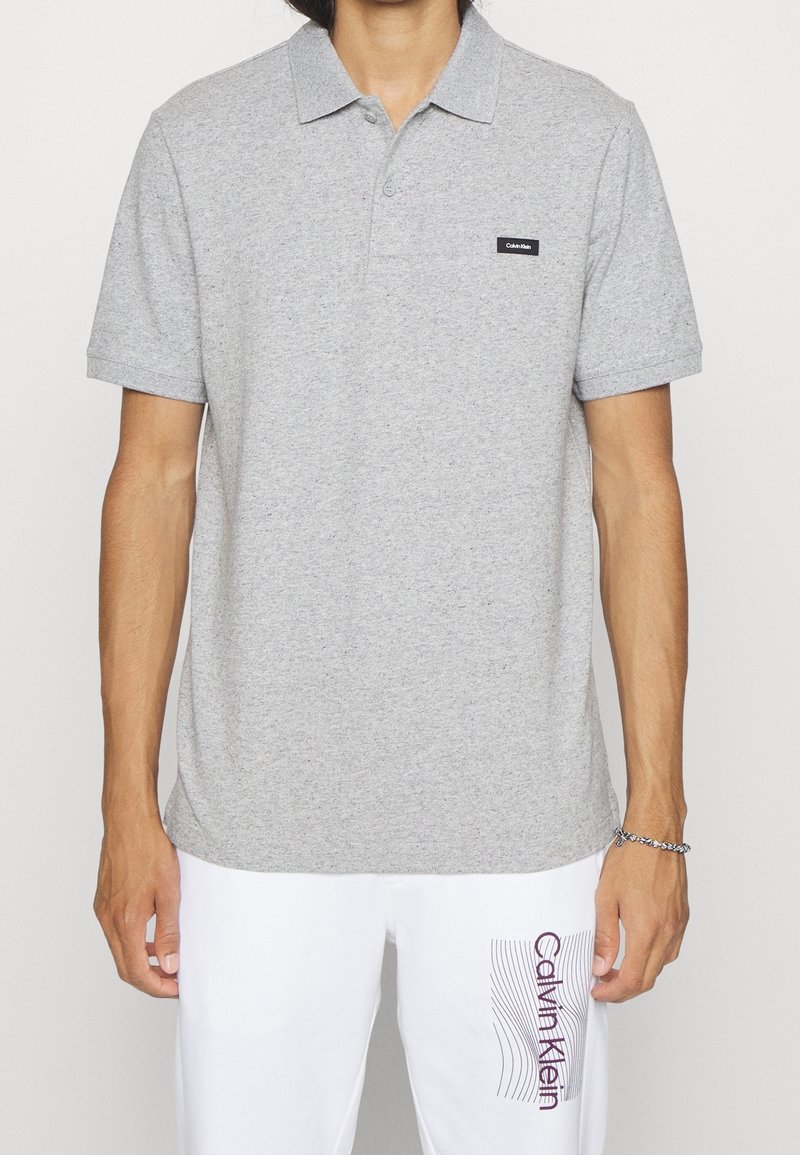 Un homme porte un polo manches courtes gris clair avec un petit logo noir Calvin Klein et un pantalon blanc avec le texte Calvin Klein sur la cuisse gauche.