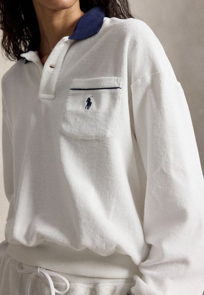 Nő fehér, hosszú ujjú galléros pólót visel, tengerészkék gallérral és mellzsebbel, amelyen egy kis hímzett ló logó látható.