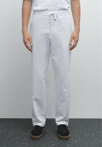 Witte katoenen broeken hebben een elastische tailleband met een trekkoord. De pasvorm is recht en ze zijn gecombineerd met donkere schoenen op een gestructureerd oppervlak.