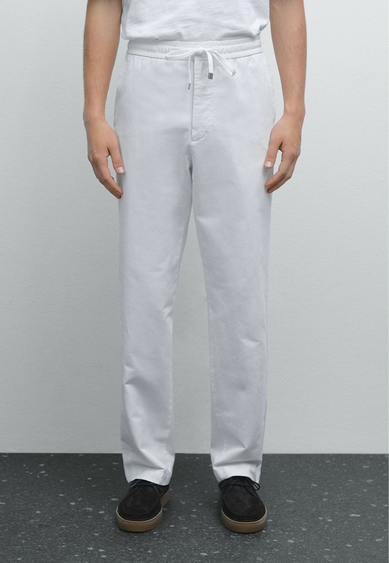 Witte katoenen broeken hebben een elastische tailleband met een trekkoord. De pasvorm is recht en ze zijn gecombineerd met donkere schoenen op een gestructureerd oppervlak.