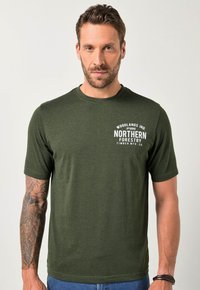 Muž s krátkými hnědými vlasy a vousy, oblečený v tmavozeleném tričku "Northern Forestry", ukazující podrobný tetování na levém paži na jednobodovém pozadí.
