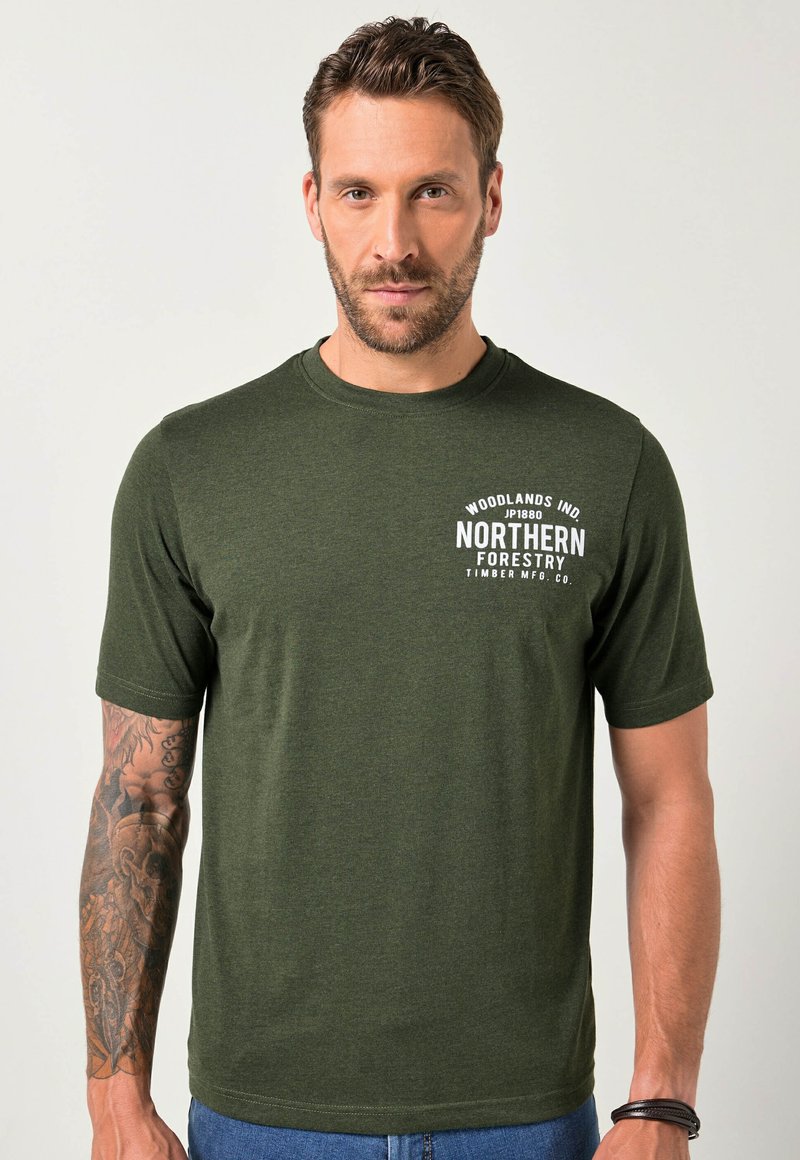 Muž s krátkými hnědými vlasy a vousy, oblečený v tmavozeleném tričku "Northern Forestry", ukazující podrobný tetování na levém paži na jednobodovém pozadí.