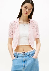 Cardigan de punto rosa de manga corta con cuello, que presenta un diseño de calados y cierre de botones, usado sobre una blusa blanca de fruncido y shorts de mezclilla.