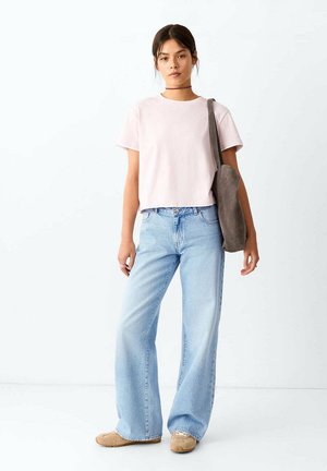 Jeune femme portant un t-shirt rose clair, un jean bleu clair à jambes larges, des chaussures marron clair, un collier noir, portant un grand sac d'épaule gris, debout sur fond blanc.