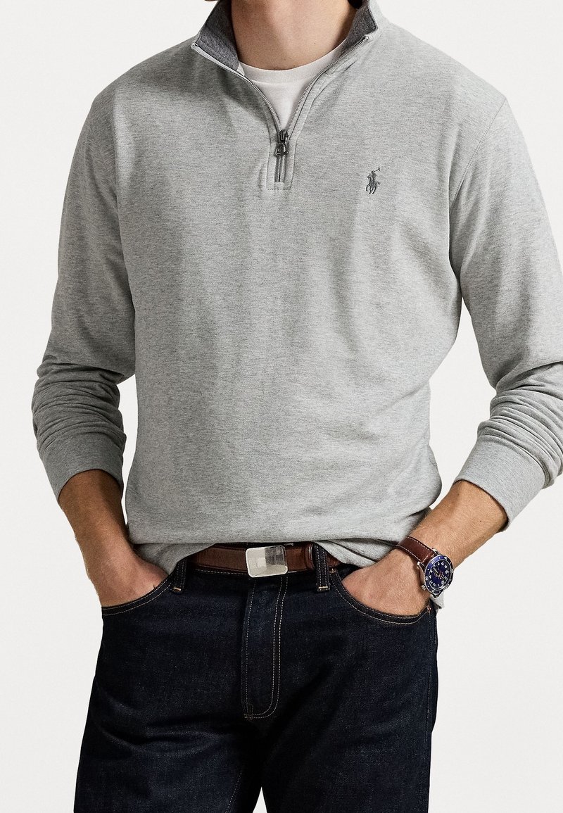 Homme portant un sweat-shirt gris clair à fermeture éclair quart, avec petit logo brodé, jean foncé, ceinture en cuir marron et montre-bracelet à cadran bleu.